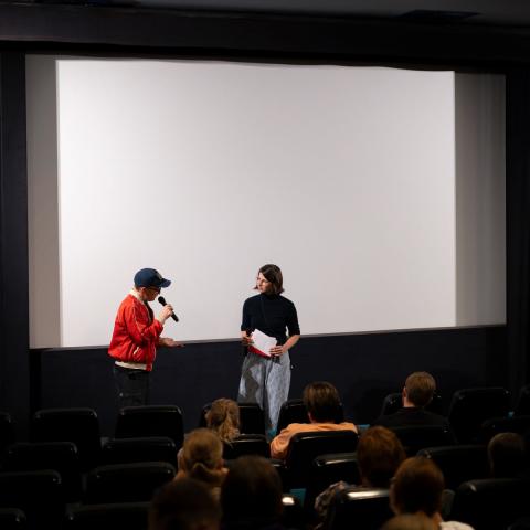Tina Jany, DOK Bildung Koordinatorin im Filmgespräch mit einer Regisseurin in einem Kinosaal voller Kinder im Publikum