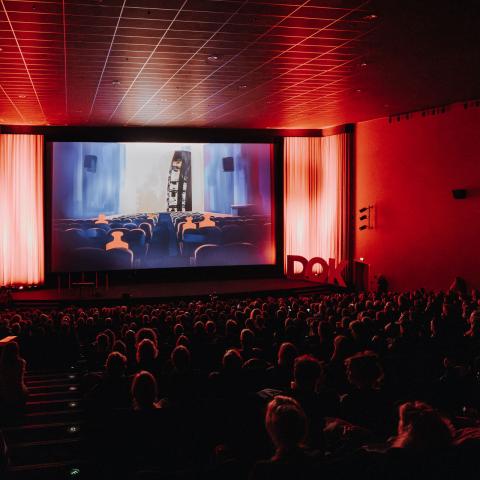 A fully occupied cinema hall, lit up in atmospheric red light. An excerpt from the festival trailer can be seen on the screen: small red weights, also sitting in a cinema.