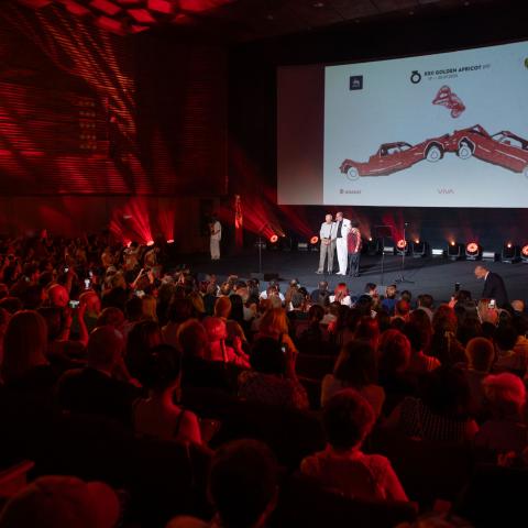 Audience in a cinema hall looks toward a stage with three people. The screen shows the poster design of GIAFF 2025.
