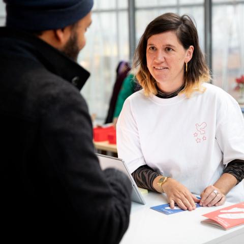 Eine Volunteer steht lächelnd am Gästecounter und unterhält sich mit einem DOK Leipzig Besucher.