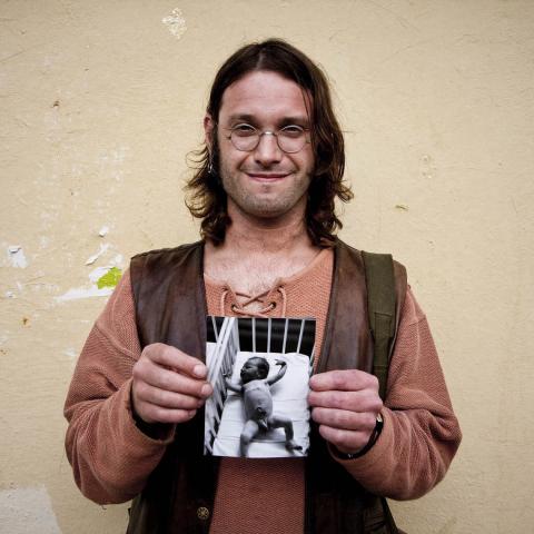 A man with glasses and long brown hair is holding a black-and-white photograph of a baby in both hands.