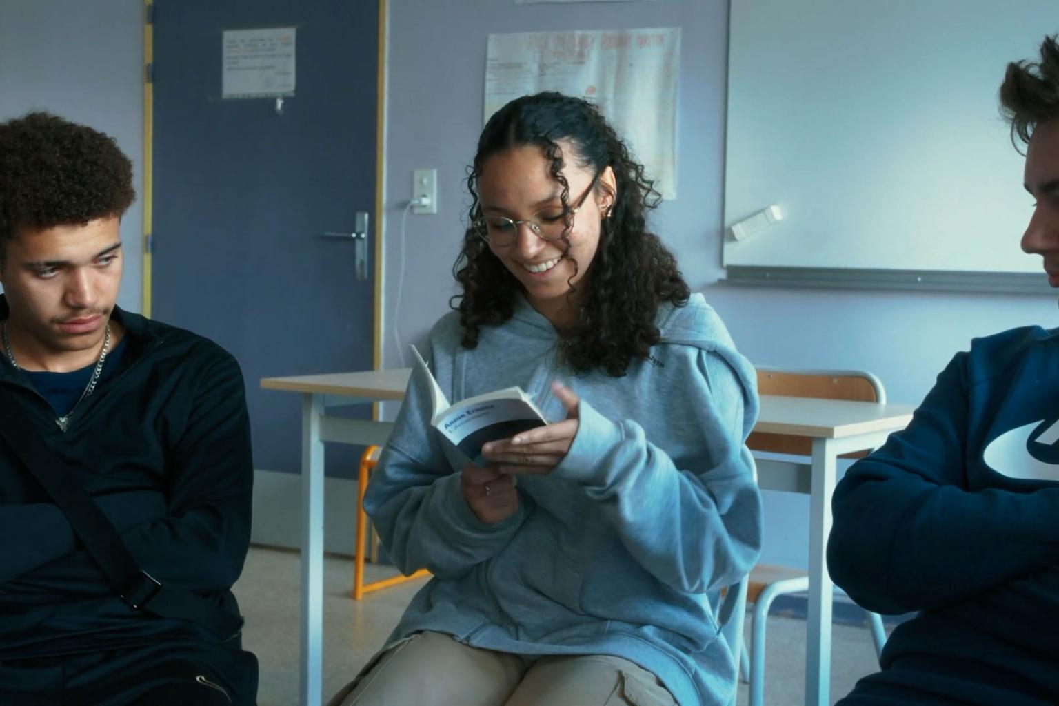 Three teenagers in a classroom, two look quite bored but the one in the middle is holding a small book in her hands and reads from it, grinning