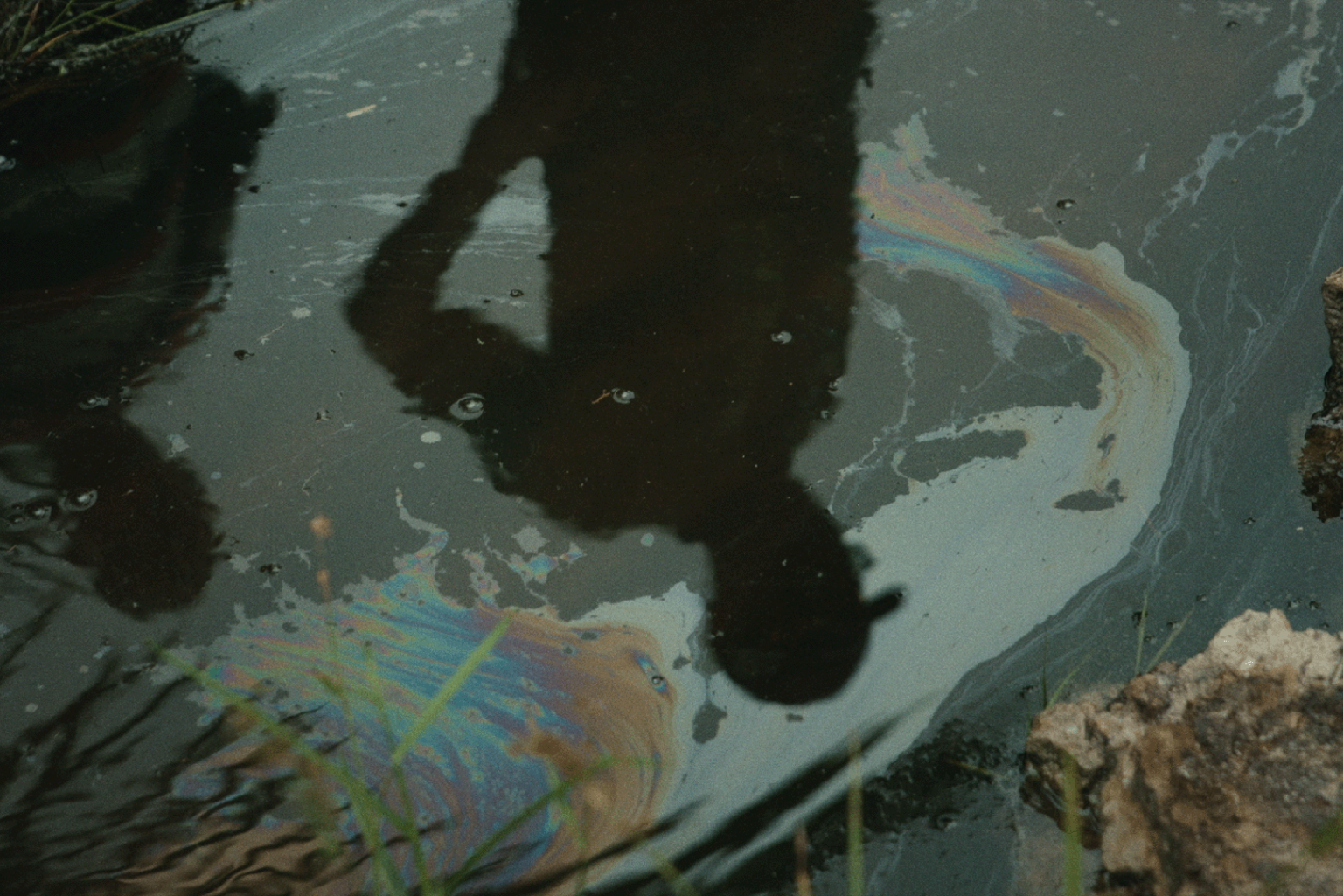 On the surface of oily rainbow-coloured water the silhouette of a person looking down can be seen