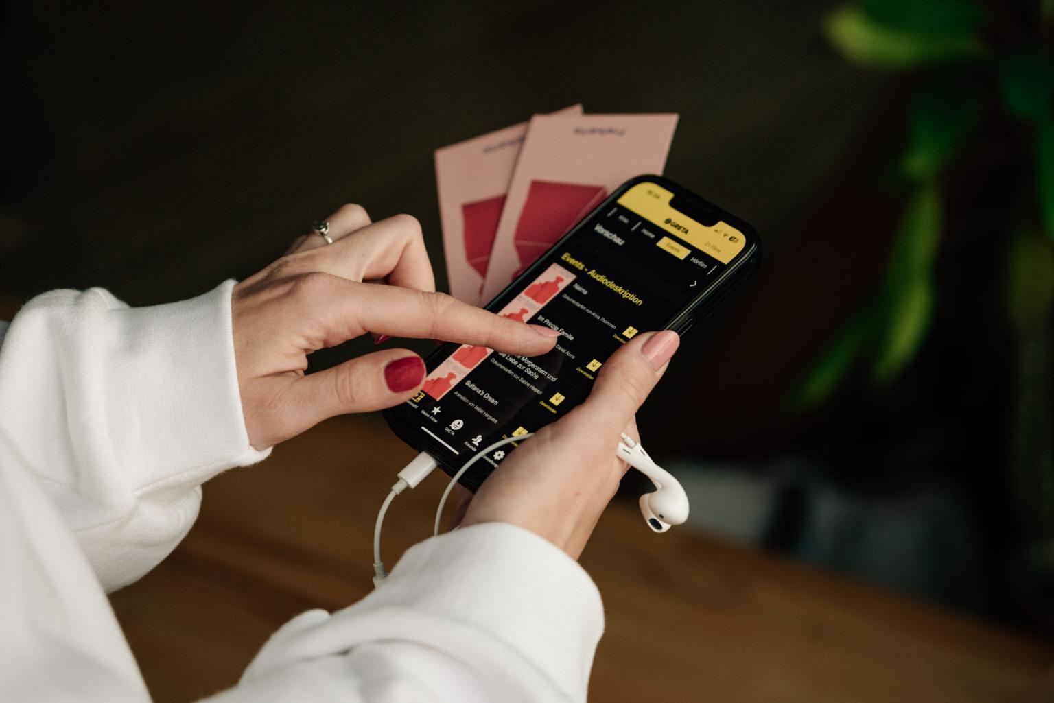 Nahaufnahme von Händen, die ein Handy halten, auf dem die Greta App für Audiodeskriptionen geöffnet ist