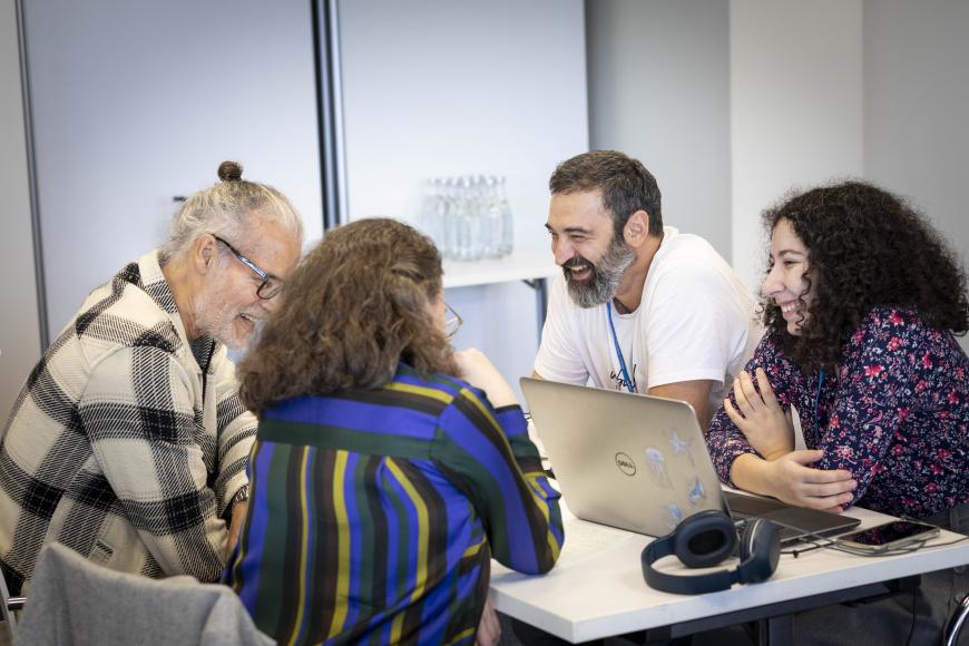Four people at a table are all laughing in conversation. Between them is a laptop, headphones and notebooks on the table.