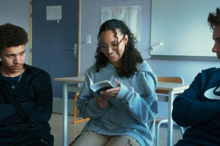 In einem Klassenraum sitzen drei Teenager, das Mädchen in der Mitte hält ein dünnes Buch in den Händen und liest grinsend daraus vor
