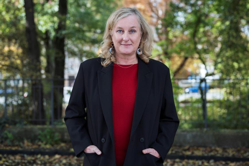 Eine Person mit blonden, schulterlangen Haaren steht im Freien vor Bäumen, trägt einen schwarzen Blazer über einem roten Oberteil und blickt leicht lächelnd direkt in die Kamera.