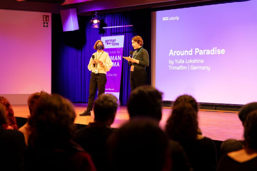 Zwei Personen auf der Bühne, die linke trägt eine Maske. Vor ihnen mehrere Leute aus dem Publikum zu sehen und hinter ihnen der Text "Around Paradise by Yulia Lokshina"